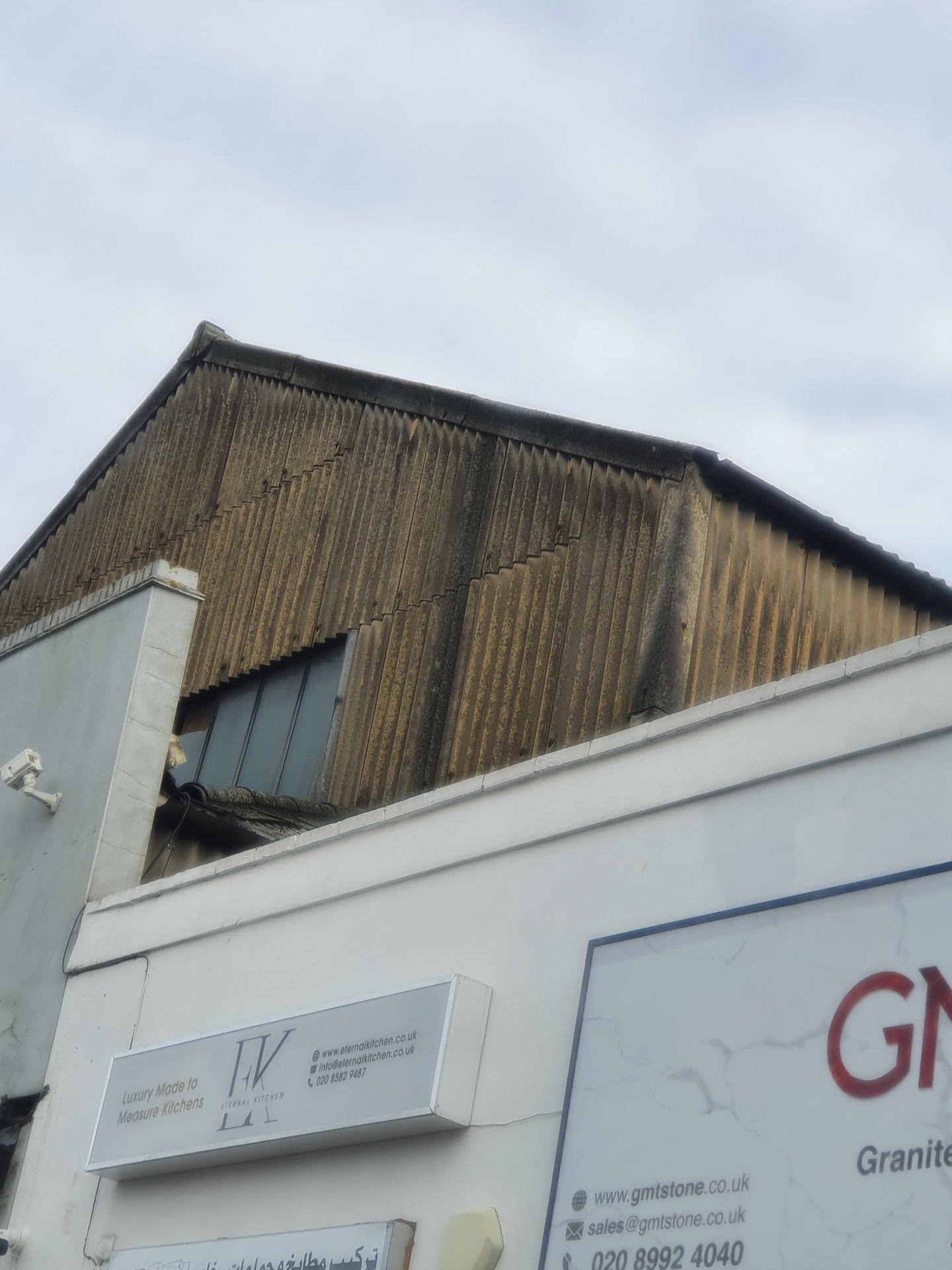 Weathered corrugated asbestos cement roof on a London commercial building with surface erosion.