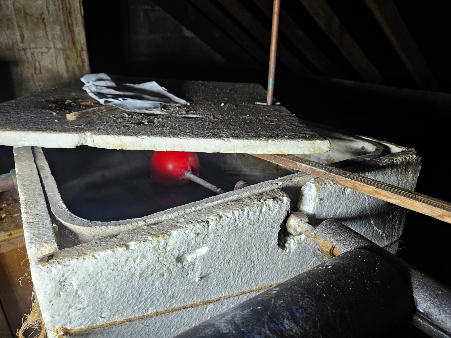 Open asbestos cement cold water cistern in a loft with red ball-valve float visible and a timber board lid lifted.
