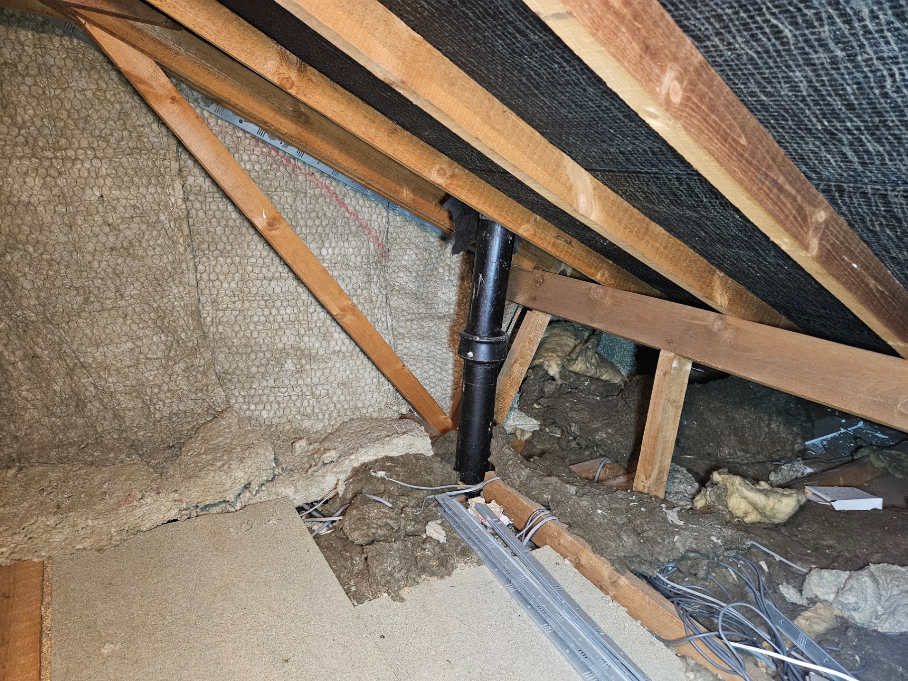 Brown fibrous lagging wrapped around an old soil pipe in a loft, surrounded by loose mineral wool insulation between rafters.