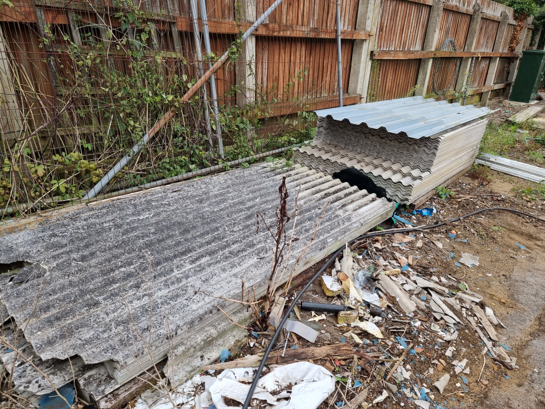 Stacked corrugated asbestos cement roofing sheets stored externally beside a fence in a yard.
