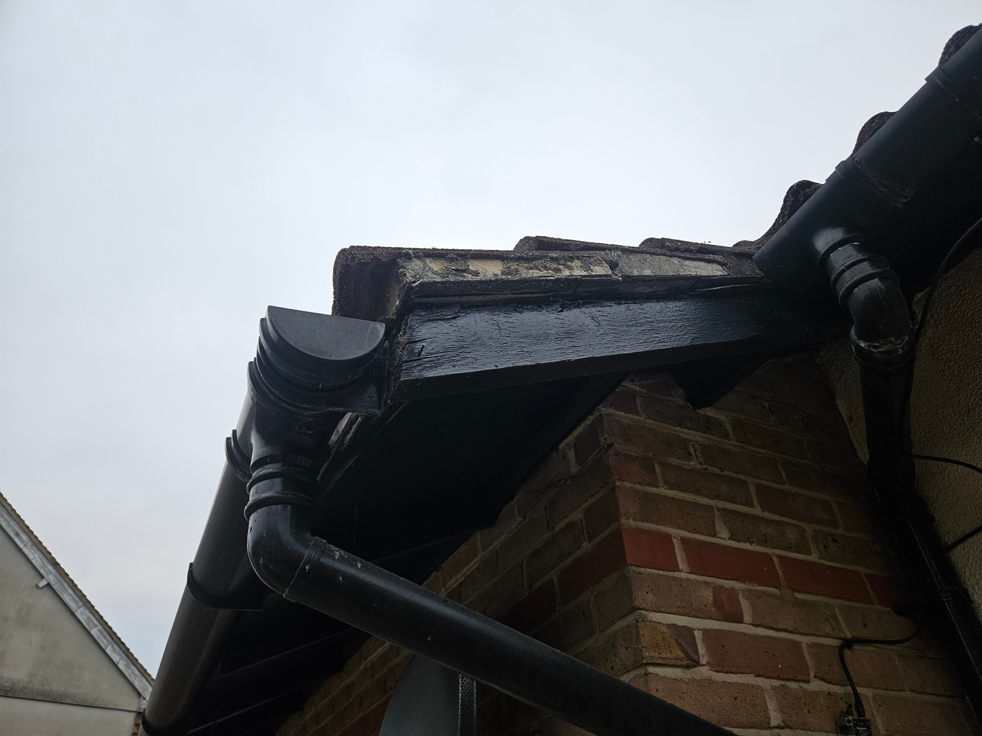 Damaged soffit or eaves board at a roof edge beside black rainwater goods, with exposed weathered board material visible along the return.