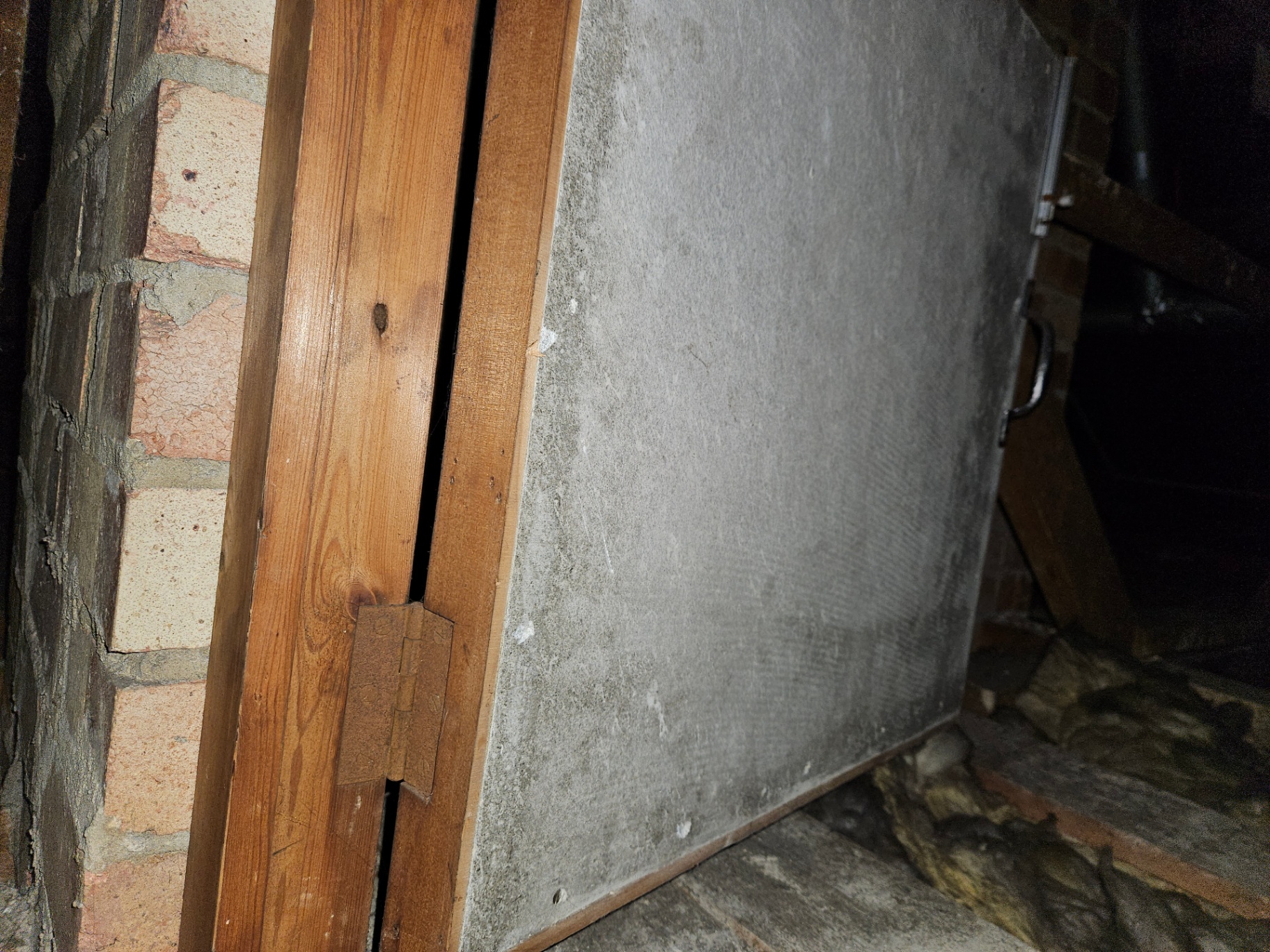 Grey asbestos insulating board fixed to a timber frame forming a small cupboard door beside a brick chimney.
