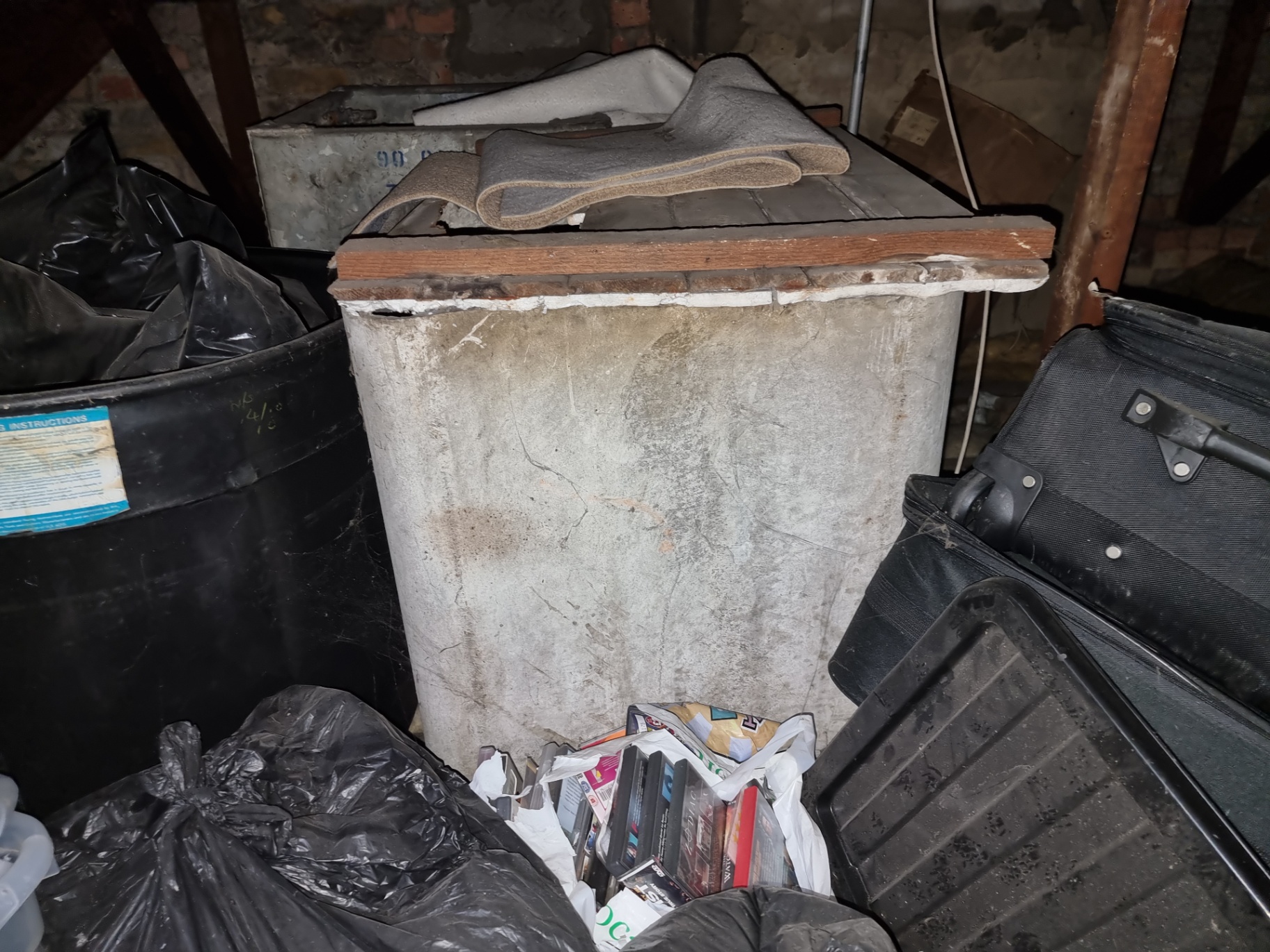 Old asbestos cement cold water tank stored in a loft among household items and bags.