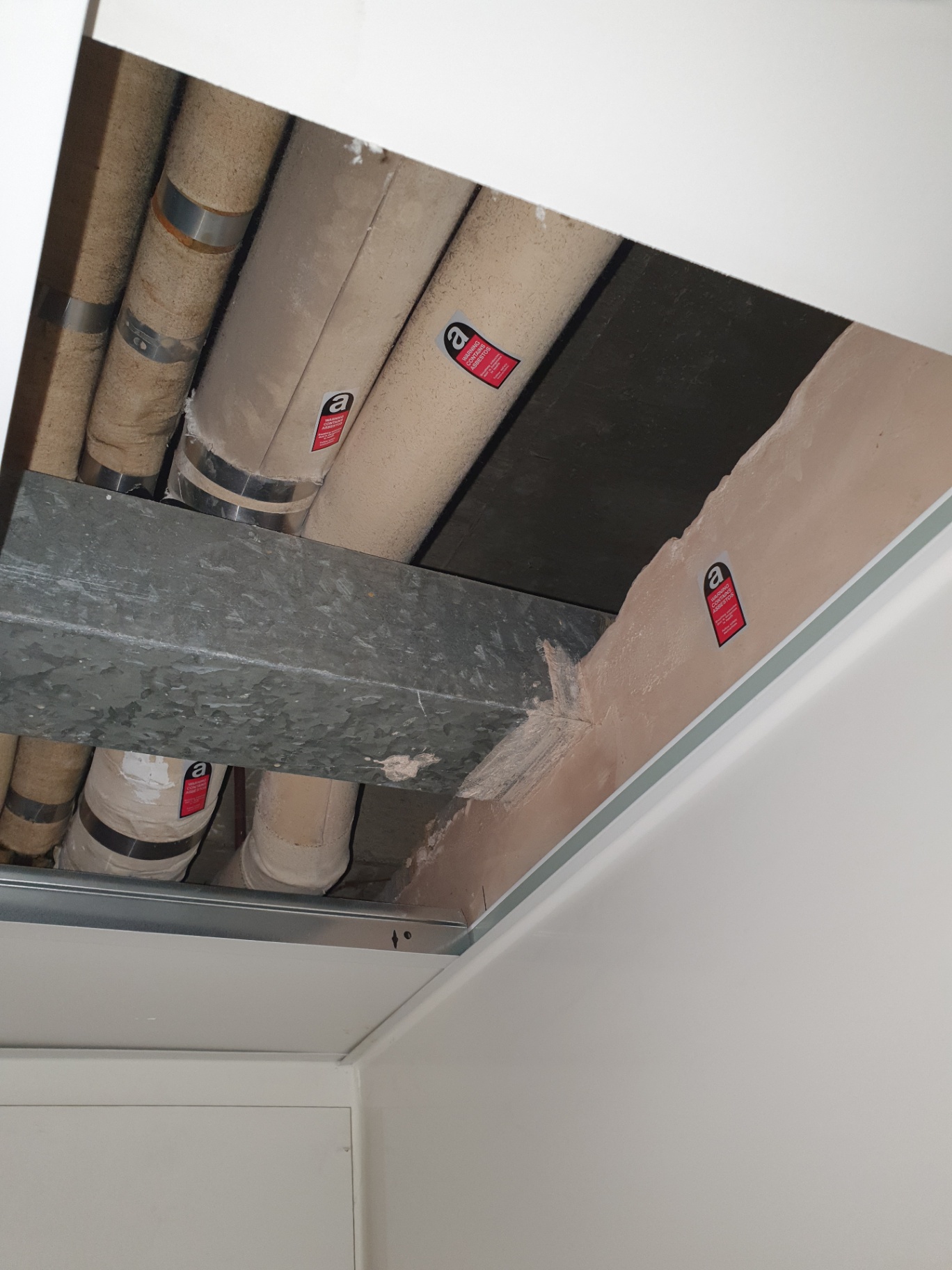 Asbestos pipe lagging with warning labels exposed above a removed ceiling tile in a commercial property.
