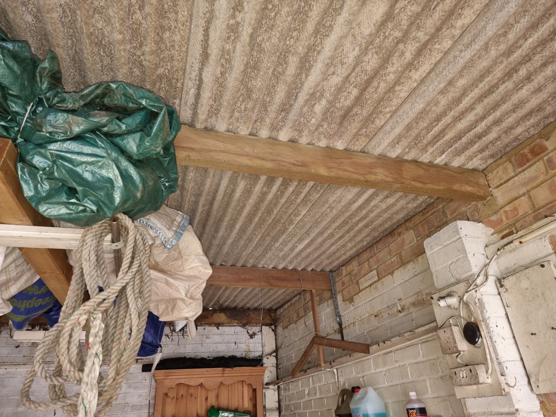 Internal view of weathered corrugated asbestos cement roof sheets supported by timber beams in a garage.